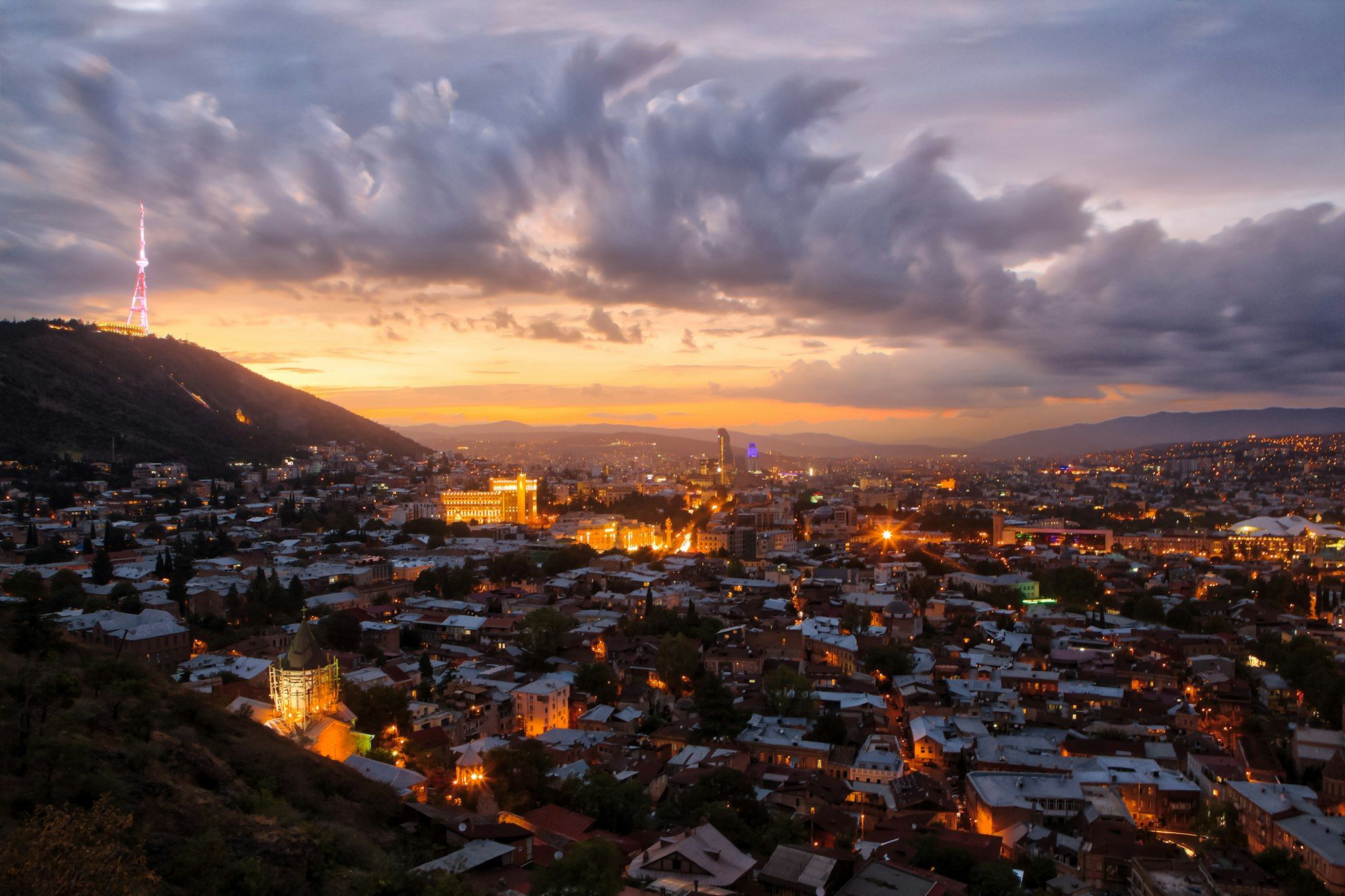 Old Tbilisi rooftops and Mtatsminda hill under a pink and gold dusk, where the honeymoon begins