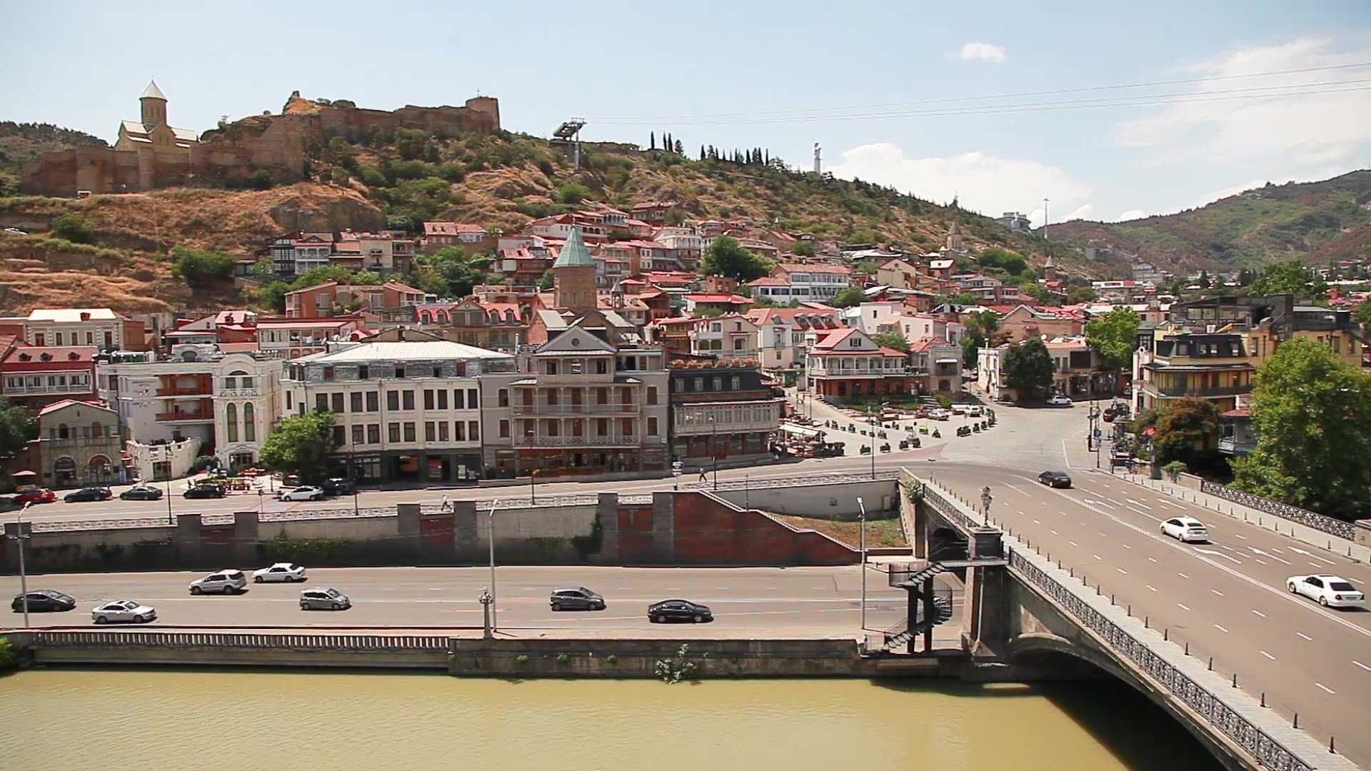 Old Tbilisi from above: Narikala fortress on its ridge, the Metekhi Bridge over the Kura, and the balconied houses of the old town in the summer sun